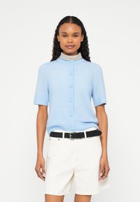 Gaiši zils īsroku kardigans ar apaļu apkakli un pogu aizdari. Mīksta tekstūra, valkāts kopā ar baltām šortiem un melnu jostu.