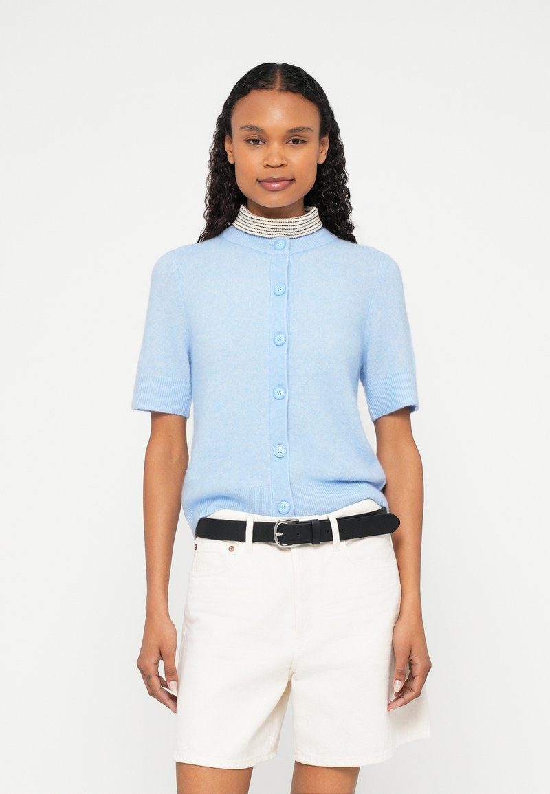 Gaiši zils īsroku kardigans ar apaļu apkakli un pogu aizdari. Mīksta tekstūra, valkāts kopā ar baltām šortiem un melnu jostu.