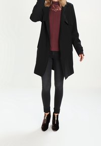 Cappotto lungo nero con apertura frontale, design a strati, indossato sopra una camicia bordeaux con pizzo, abbinato a jeans skinny scuri e stivaletti.