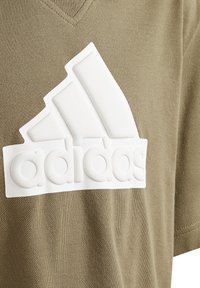 Olivengroen T-shirt gemaakt van gestructureerde stof met een groot wit rubberen logo en drie horizontale strepen op de borst.