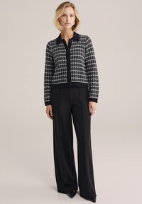 Čiernobiely károvaný kardigán s golierom, prednými gombíkmi a rebrovanými manžetami, skombinovaný s vysokými čiernymi nohavicami a špicatými podpätkami.
