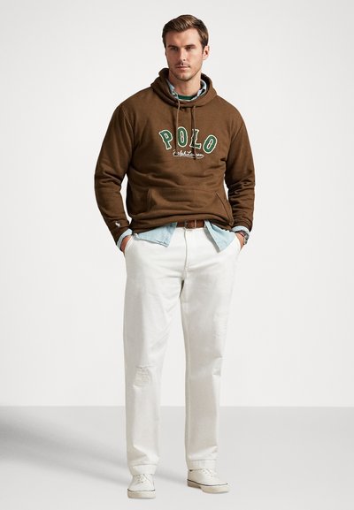 Άνδρας που φοράει καφέ φούτερ "Polo" πάνω από ανοιχτό μπλε πουκάμισο, λευκό παντελόνι, λευκά αθλητικά παπούτσια, στέκεται με τα χέρια στις τσέπες μπροστά σε απλό φόντο.