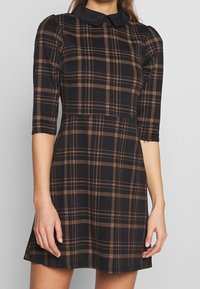 Svart rutig klänning med bruna och beiga linjer, med svart krage, trekvartsärmar och en utsvängd kjoldesign.