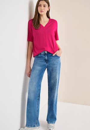 Frau, die vor einer Wand steht, trägt ein hellrosa V-Ausschnitt-Oberteil, hellblaue Jeans mit weitem Bein und silberne Sneakers, eine Hand in der Tasche.