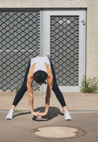 Sportieve outfit met een mouwloos wit topje en zwarte leggings. De figuur strekt zich naar voren op een verharde ondergrond, met een grijze metalen rooster op de achtergrond.