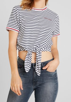Pasiasty krótki top, biały z granatowymi poziomymi paskami, czerwone wykończenie na rękawach i dekolcie, zawiązany węzłem z przodu, w połączeniu z ciemnymi dżinsami.