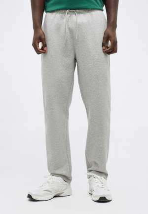 Isik kannab heledaid halli värvi dressipükse, millel on nöör, ja valgeid spordijalatseid, seisab ühevärvilise tausta ees.