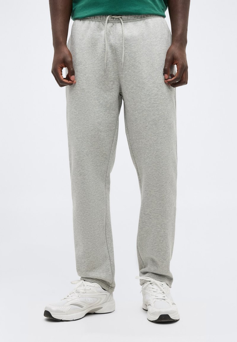 Άτομο που φοράει ανοιχτό γκρι φόρμα με κορδόνι και λευκά αθλητικά παπούτσια, στέκεται μπροστά σε απλό φόντο.