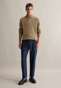 Beiger, gerippter Strickpullover mit halbem Reißverschluss und dunkeltblauen, locker sitzenden Hosen sowie schwarzen Mokassins. Einfaches Design, neutrale Farben.