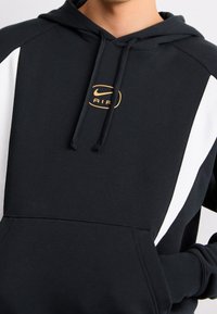 Black hoodie with white side panels, features a large gold logo on the chest and a kangaroo pocket. Made from soft, textured material.