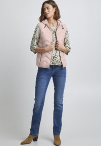 Gilet rembourré rose avec capuche, motif matelassé, porté sur une chemise légère à fleurs, associé à un jean blue et des bottines beige.