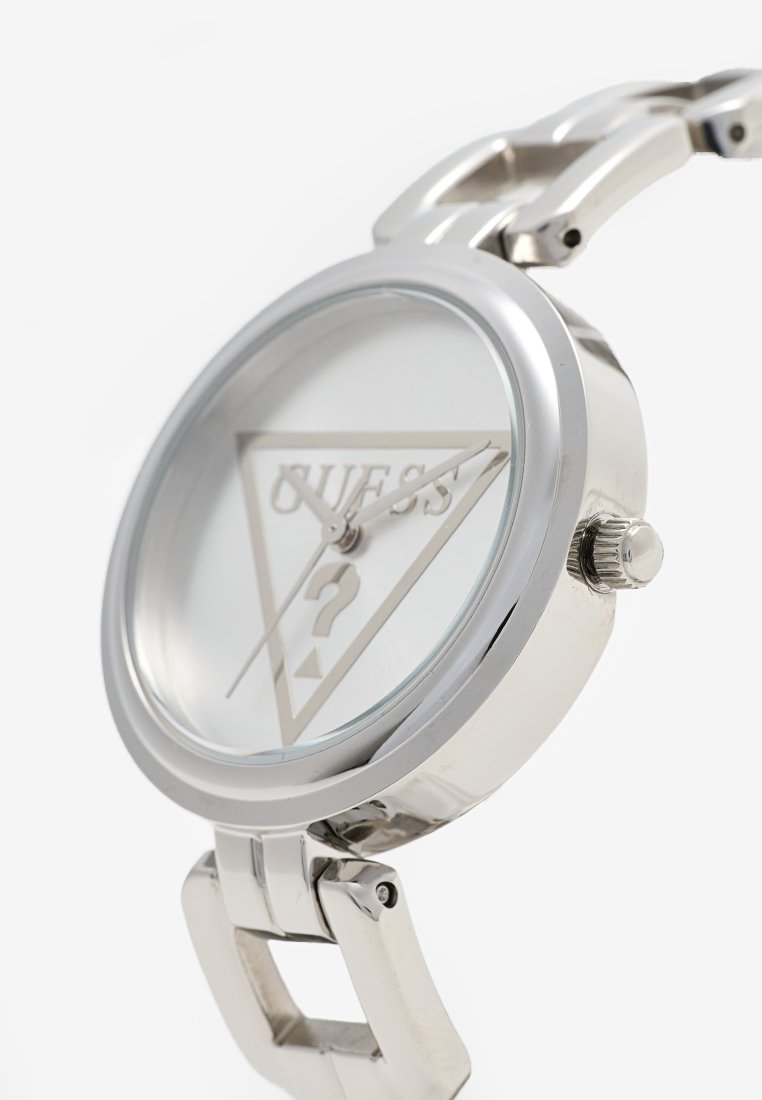 Montre en métal argenté avec un cadran rond, verre transparent et un fond blanc affichant le logo "Guess" et un point d'interrogation. Présente un bracelet élégant.
