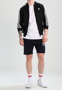 Černá zipová bunda s bílým logem Adidas, bílá tričko, námořnické šortky a bílé tenisky s červenými pruhovanými ponožkami. Neformální sportovní oblečení.