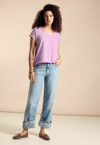 Maglietta viola a maniche corte con scollo a V, jeans azzurri a gamba larga con orli risvoltati e scarpe basse marroni. Tessuto morbido, vestibilità rilassata.