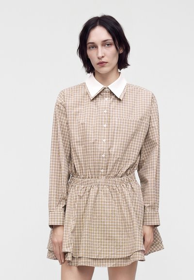 Jeune femme aux cheveux courts et foncés porte une robe à manches longues beige à carreaux avec un col blanc et une taille froncée sur un fond uni.