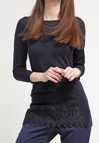 Marinblå långärmad topp med en texturerad svart spetskant, med en rund halsringning och figurnära silhuett. Slätt stickat tyg.