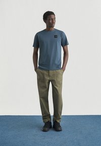 Ανδρικό μπλε σκούρο μπλουζάκι με μαύρο λογότυπο, συνδυασμένο με χακί παντελόνι. Μαύρα παπούτσια; απλό φόντο με μπλε δάπεδο.