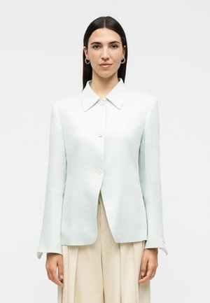 Woman with straight dark hair wearing a light blue buttoned blazer and beige pleated pants, standing against a plain light background.