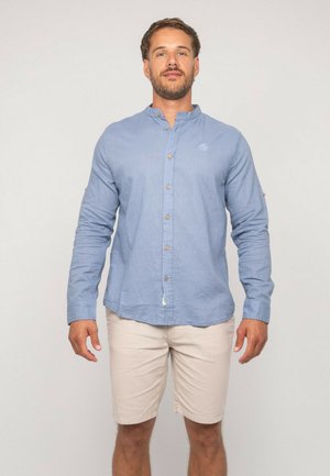 Homme portant une chemise à manches longues bleu clair à boutons avec col mandarin et un short beige, debout devant un fond clair uni.
