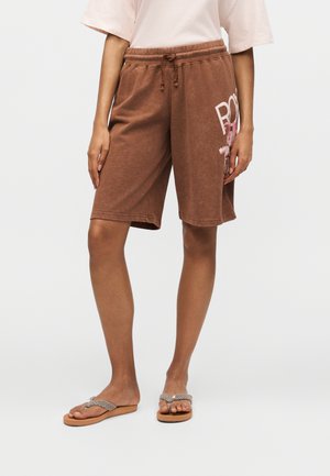 Persona que lleva puestos pantalones cortos marrones hasta la rodilla con cordón y un gráfico en la pierna derecha, camiseta beige y chanclas con tiras trenzadas.