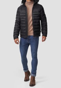 Veste noire matelassée avec fermeture éclair et capuche, associée à un col roulé beige, un jean bleu et des bottines en cuir marron.