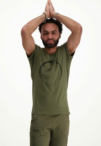 Oliivinvihreä T-paita, jossa on musta pyöreä kuvio edessä, lyhyet hihat ja pyöreä kaula-aukko, yhdistettynä samanlaisiin oliivinvihreisiin housuihin.