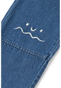 Blauer Denim-Stoff mit einem weißen bestickten Design aus wavy Linien, dazu zwei Punkte und ein Lächeln, positioniert im Taschenbereich.
