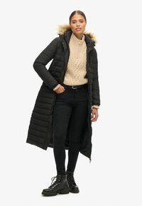 Abrigo negro largo y acolchado con capucha de piel sintética desmontable, combinado con un suéter de punto con trenzas crema y jeans negros. Botas negras de cordones completan el look.