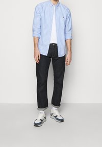 Jasnoniebieska i biała koszula z rozpiętymi guzikami noszona na białej koszulce, w zestawieniu z ciemnoniebieskimi dżinsami i wielokolorowymi trampkami.