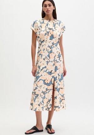 Femme portant une robe midi sans manches à grand imprimé floral pêche et bleu, fente à l'avant, et sandales plates noires sur un fond uni.