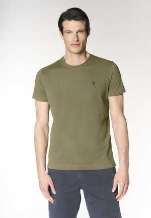 Uomo che indossa una t-shirt verde oliva a maniche corte con scollo rotondo e pantaloni blu navy, in piedi davanti a uno sfondo chiaro, rivolto in avanti.