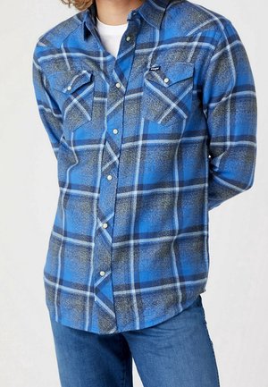 Person trägt ein blau-schwarz-weiß kariertes Hemd über einem weißen Unterhemd, kombiniert mit blauen Jeans, vor einem neutralen Hintergrund.