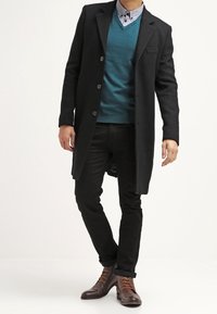 Černý vlněný kabát s dlouhým střihem, pod ním tyrkysový svetr, šedá košile s knoflíky, černé kalhoty a hnědé šněrovací boty.