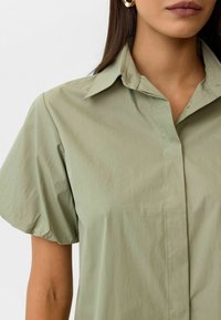 Licht olijfgroene shirt met korte mouwen, klassieke kraag, fijne textuur en details van een knoopsluiting aan de voorkant. Licht oversized pasvorm.