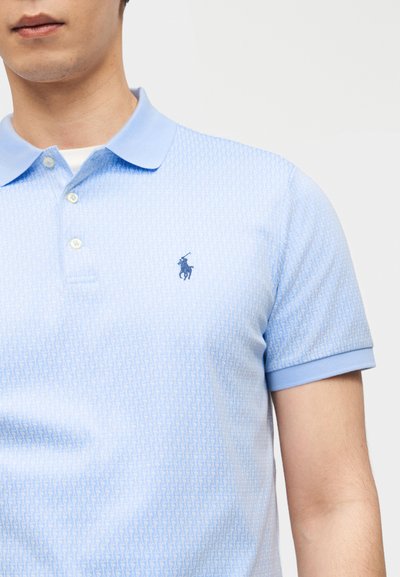 Άνδρας που φοράει ανοιχτομπλε πόλο πουκάμισο με διακριτικό μοτίβο και σκούρο μπλε κεντημένο λογότυπο παίκτη πόλο στο στήθος.