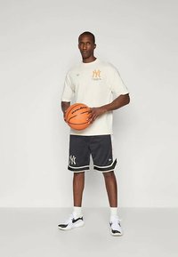 Ανοιχτό μπεζ μπλουζάκι με πορτοκαλί λογότυπο, μαύρες αθλητικές βερμούδες με λευκό περίγραμμα και λογότυπο, λευκές κάλτσες και μαύρα αθλητικά παπούτσια, κρατώντας μια μπάλα μπάσκετ.