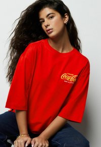 Czerwona bawełniana koszulka o luźnym kroju i krótkich rękawach, z żółtym haftowanym logo Coca-Cola na lewej stronie klatki piersiowej.