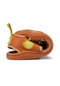 Zapato de cuero marrón con una textura suave, forma redondeada y acentos amarillos. Logotipo exhibido en el lateral; incluye una pestaña de agarre para facilitar el uso.