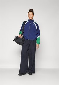 Giacca a vento blu navy e verde con dettagli bianchi, dotata di colletto alto e chiusura con zip, abbinata a pantaloni larghi scuri e una borsa nera.
