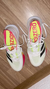 White and neon Adidas Adizero running shoes with black stripes, white laces, and bright yellow insoles, placed on a wooden floor.