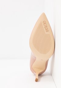 Chaussure beige à talon haut à bout pointu reposant sur le côté montrant la semelle texturée avec le logo "GUESS" et la taille 38.