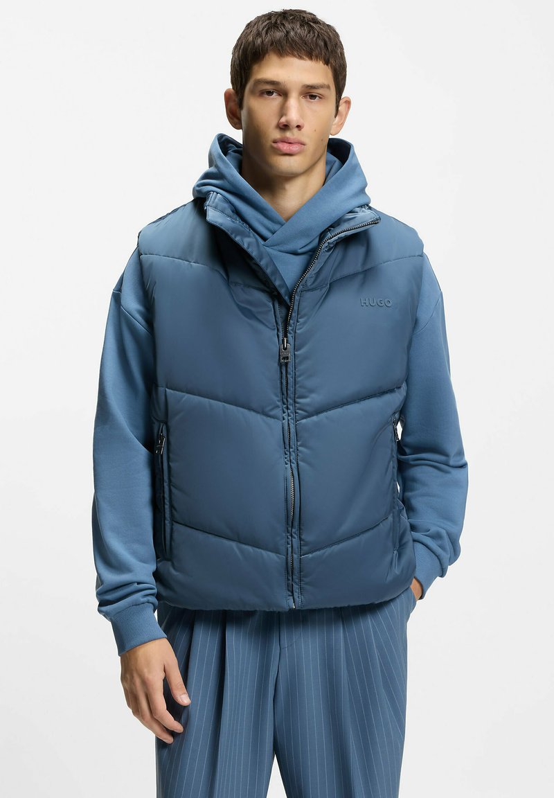 Jeune homme portant une veste matelassée bleue par-dessus un sweat à capuche bleu et un pantalon bleu à fines rayures, debout avec une main dans la poche.