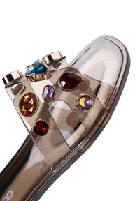 Sandale transparente à bout rond, ornée d'accents en bijoux multicolores. Surface lisse et brillante avec un léger talon.