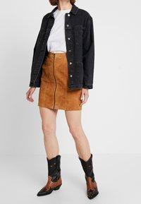 Giacca di jeans nera sopra una maglietta bianca, abbinata a una gonna in suede beige con zip e stivali da cowboy neri con dettagli marroni e stelle.