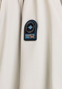 Lichtbeige jassenstof met een zachte textuur. Bevat een donkerblauwe patch met "Established 1955" en een logo in zwarte en aqua accenten.