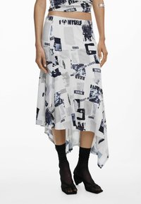Asymmetrischer Rock aus weißem Stoff, der eine Collage aus schwarzen und navyblauen Drucken mit Text und Bildern zeigt. Kombiniert mit schwarzen Socken und Absatzsandalen.