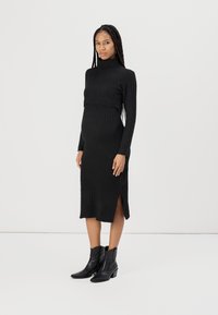 Abito a maglia nera a coste con collo alto, maniche lunghe, spacchi laterali, silhouette aderente, abbinato a stivali cowboy neri.