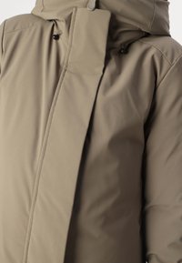 Khaki foret jakke med høj krave, synlige syninger og hætte. Har knapper og tryklåse, lavet af et glat, mat stof.