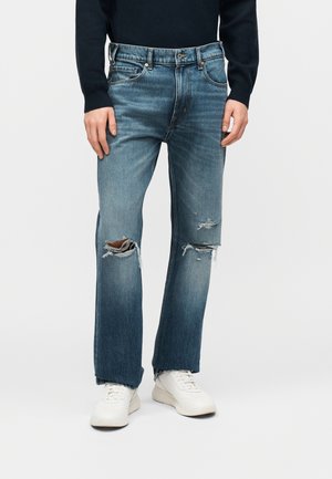 Személy közép kék, kopott farmernadrágot visel, amelynek a térdnél szakadt részei vannak, fehér sportcipőt és sötétkék, hosszú ujjú felsőt, egyszerű háttér előtt áll.