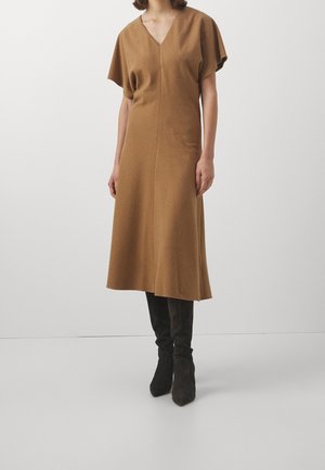 Woman wearing a brown V-neck midi dress with short sleeves paired with black knee-high boots, standing against a plain light background.
